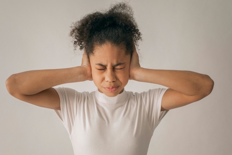 Young person with eyes closed pressing both hands over their ears, appearing distressed or trying to block out noise, against a plain background.