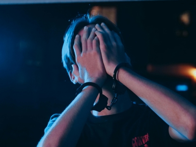 Person in handcuffs covering their face with both hands, illuminated by blue and red lighting, suggesting distress or arrest.