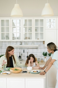 Two adults and a child gathered around a kitchen island, with plates of food and milk on the counter, appearing to have a quiet or tense moment during a meal.