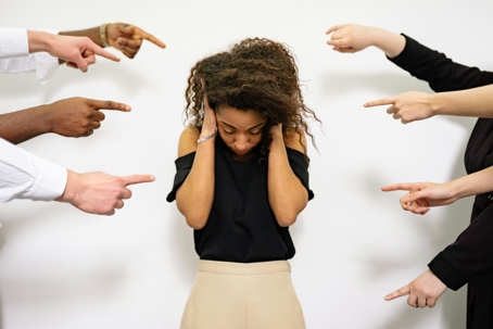 Distressed woman covering her ears while multiple hands point at her, suggesting blame, criticism, or bullying.